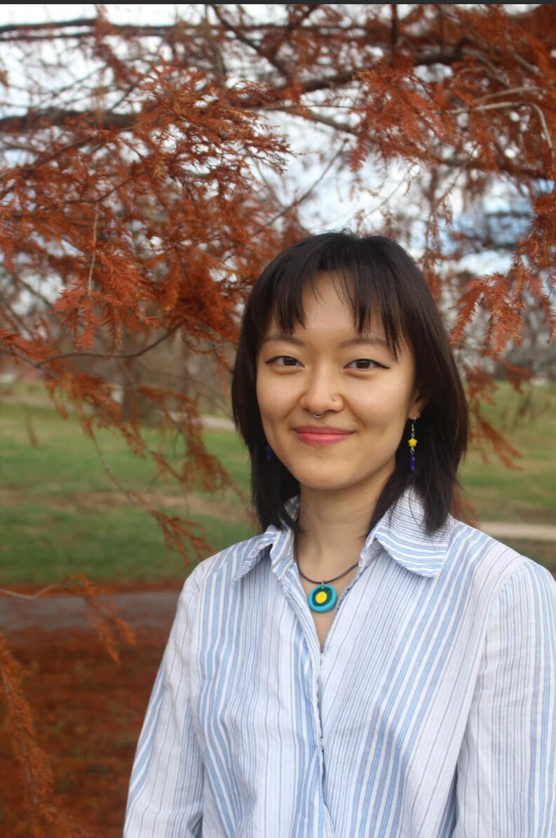 picture of an asian person in their 20's with shoulder-length black hair wearing a suit in greenery